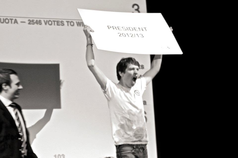 New EUSA president, James McAsh lifts a placard reading "President 2012/13" over his head. Outgoing president Matt McPherson is visible to the left of the shot.