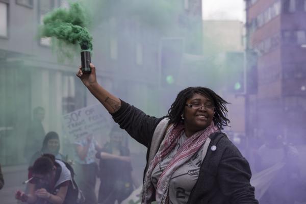 A woman of colour activist smiles whilst holding up a green smoke bomb. A crowd is visible behind them. They are attending the Sisters Uncut action da.y.