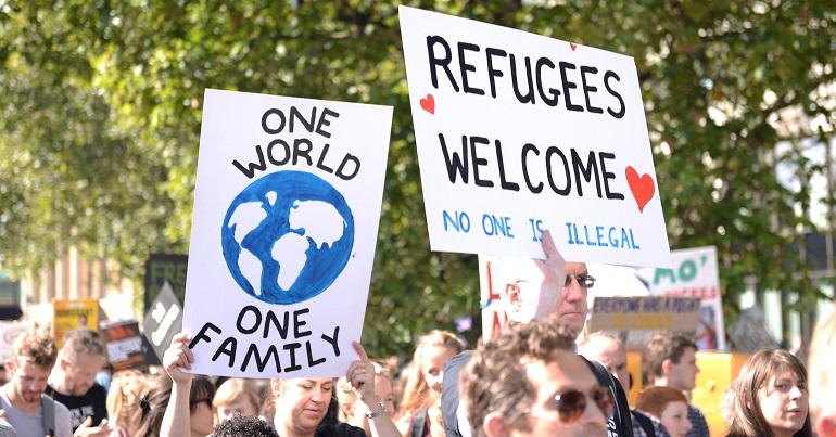 A protest with two signs, one reading "one world, one family" and the other reading "Refugees welcome, no one is illegal