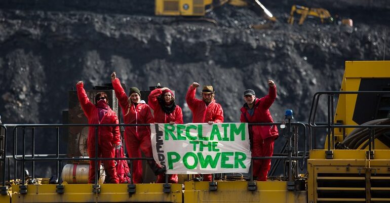 Reclaim the Power activists stood on mining equipment at an open cast coal mine, their banner reads Leeds Reclaim the Power.
