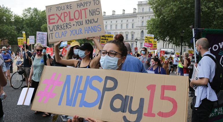 A photo of protesters with placards reading #NHSPay15 and "if youre happy to exploit NHS workers and you know it clap your hands"