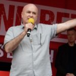 Mick Lynch speaking at a demonstration, with the words "Enough is Enough" behind him