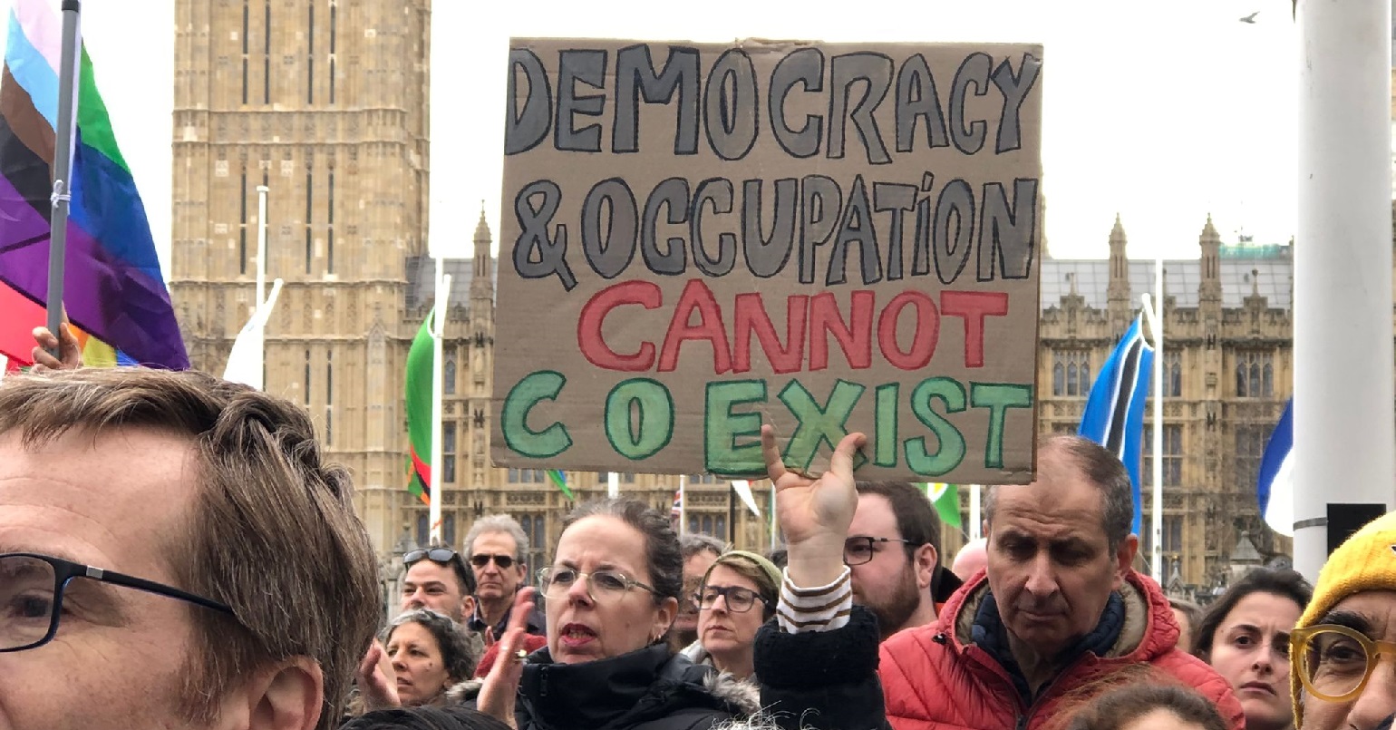 A photo of a protest in Westminster. One protester is holding a sign reading "democracy and occupation cannot co-exist"