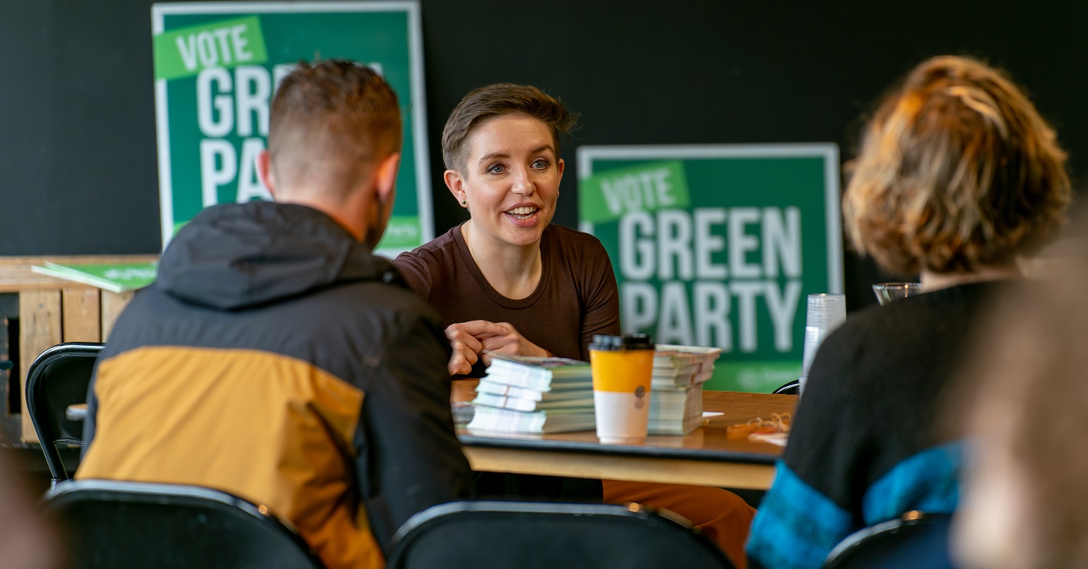 Green Party co-leader Carla Denyer sitting at a table talking with two other people. Two posters behind her read "Vote Green Party"