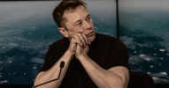 A photo of Elon Musk resting his faces on his hands in front of a microphone